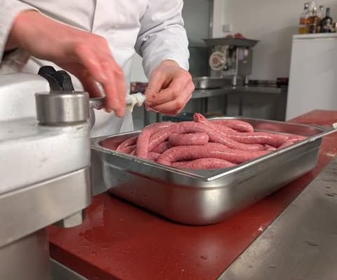 Person wearing a white coat filling raw sausages by hand into a metal tray during meat processing.